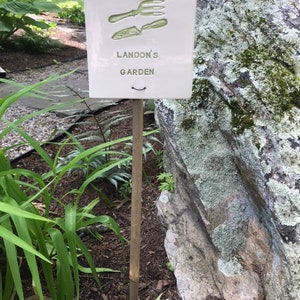 May include: A white ceramic garden sign with a green image of gardening tools and the text "Landon's Garden". The sign is attached to a wooden stake and is placed in the ground next to a large rock.