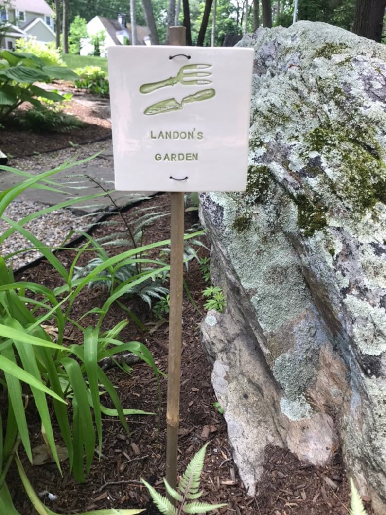 May include: A white ceramic garden sign with a green image of gardening tools and the text "Landon's Garden". The sign is attached to a wooden stake and is placed in the ground next to a large rock.