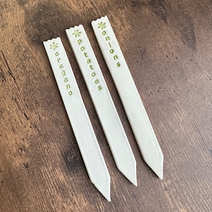 May include: Three white plant markers with green flower designs and the words "onions", "potatoes", and "oregano" printed on them.