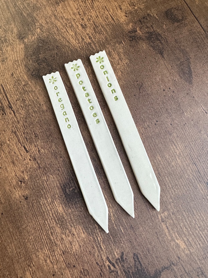 May include: Three white plant markers with green flower designs and the words "onions", "potatoes", and "oregano" printed on them.