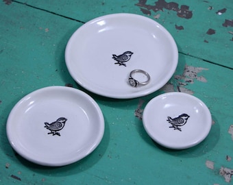 May include: Three white ceramic ring dishes with a black bird design. A silver ring with a gemstone sits on the largest dish. The dishes are arranged on a distressed teal surface.