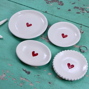May include: Five white ceramic dishes with red heart designs. The dishes are of varying sizes and shapes, and are arranged on a turquoise wooden surface.