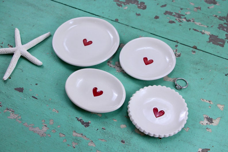 May include: Five white ceramic dishes with red heart designs. The dishes are of varying sizes and shapes, and are arranged on a turquoise wooden surface.