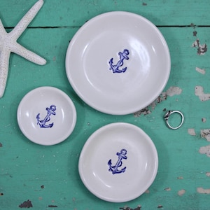 May include: Three white ceramic ring dishes with blue anchor designs. The dishes are arranged in a triangle shape on a teal wooden surface.
