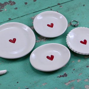 May include: Four white ceramic ring dishes with red heart designs. The dishes are of varying sizes and shapes, and are arranged on a turquoise wooden surface.