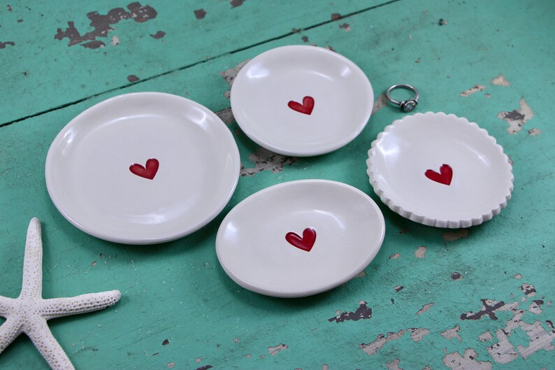 May include: Four white ceramic ring dishes with red heart designs. The dishes are of varying sizes and shapes, and are arranged on a turquoise wooden surface.
