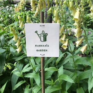 May include: A white ceramic garden marker with a green watering can illustration and the text "HANNAH'S GARDEN".