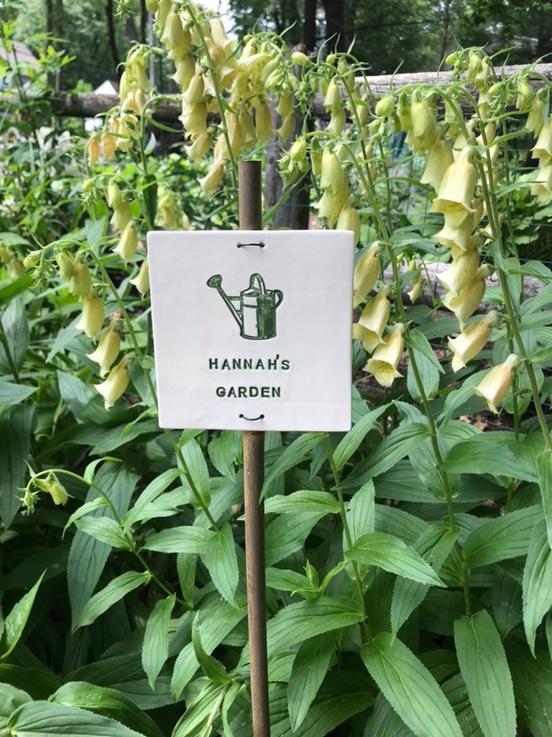 May include: A white ceramic garden marker with a green watering can illustration and the text "HANNAH'S GARDEN".