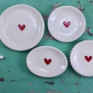 May include: Four white ceramic ring dishes with red heart designs. The dishes are on a teal background.