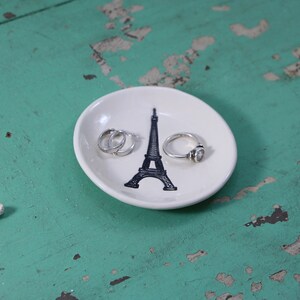 May include: White ceramic ring dish featuring a black Eiffel Tower design. Three silver rings are on the dish.