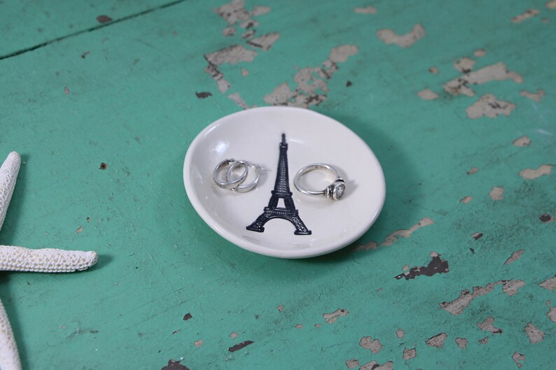 May include: White ceramic ring dish featuring a black Eiffel Tower design. Three silver rings are on the dish.