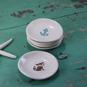 May include: Two ceramic ring dishes, one with a teal anchor design and the other with a brown fox design. The dishes are on a green wooden surface.