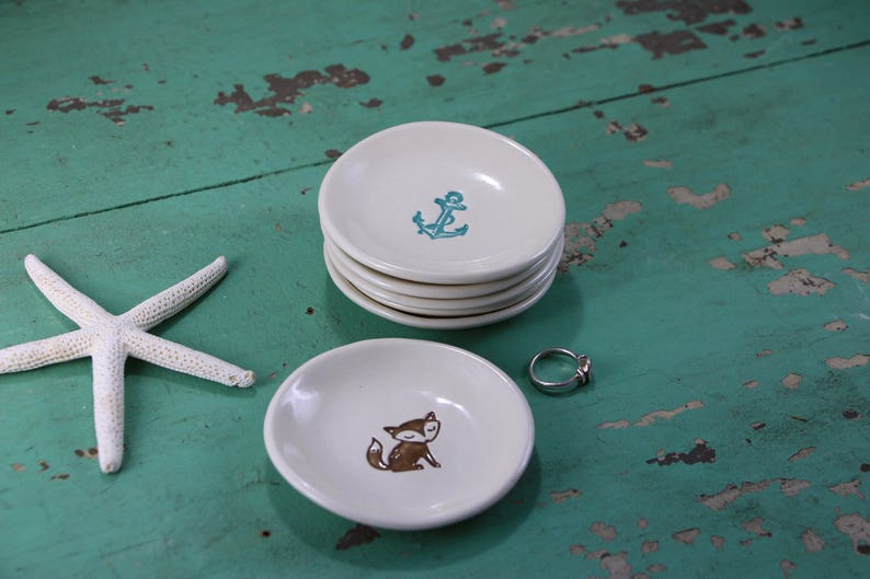 May include: Two ceramic ring dishes, one with a teal anchor design and the other with a brown fox design. The dishes are on a green wooden surface.