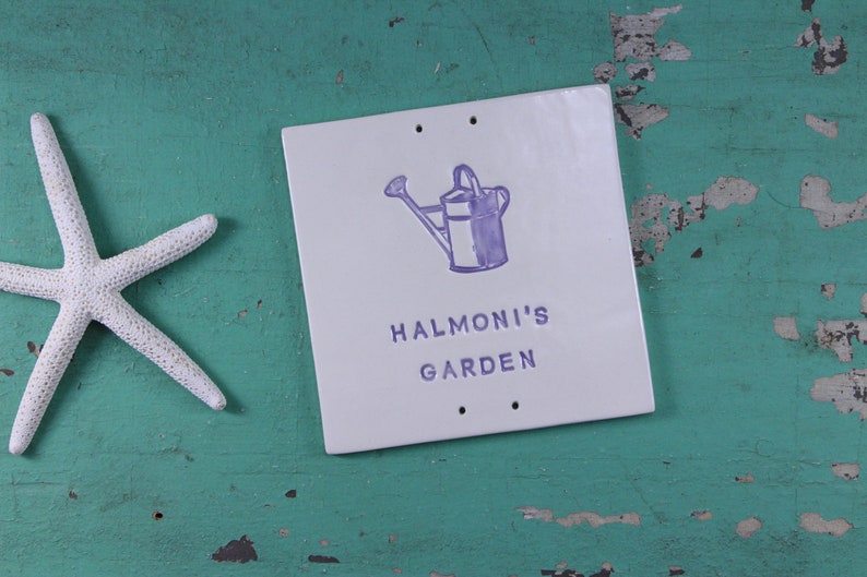 May include: A white ceramic sign with a purple watering can illustration and the text "Halmoni's Garden".