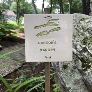 May include: A white ceramic garden marker with a green image of a trowel and a garden fork. The marker is attached to a wooden stake and reads "Landon's Garden".