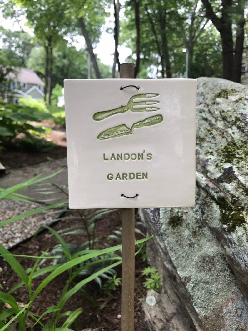 May include: A white ceramic garden marker with a green image of a trowel and a garden fork. The marker is attached to a wooden stake and reads "Landon's Garden".