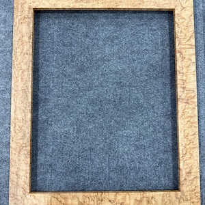 May include: A rectangular picture frame featuring a light tan wood grain with a distinct, mottled burl pattern throughout the surface. The frame has a flat, wide border design with mitered corners, showcasing the natural texture of the wood.