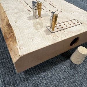 Può includere: Un tabellone da cribbage in legno con finitura in legno naturale. Il tabellone ha un bordo vivo ed è realizzato in un unico pezzo di legno. Il tabellone ha una pista di punteggio integrata e un porta-birilli. Il tabellone è mostrato con birilli in metallo e ottone.