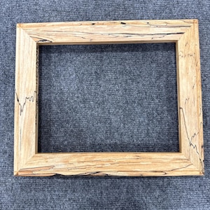 May include: A rectangular wooden picture frame with a natural wood grain finish. The frame has a light brown color with dark, irregular streaks. The frame is empty, with a visible opening in the center.
