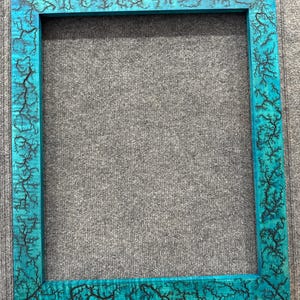 Pode incluir: Uma moldura retangular de madeira turquesa com intrincados padrões fractais pretos. O design da moldura apresenta uma estética artística única, adequada para exibir obras de arte ou fotografias. A moldura tem formato quadrado.
