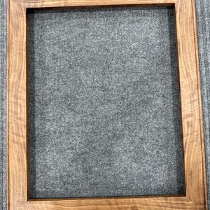 Puede incluir: Un marco de fotos rectangular hecho de madera de color marrón medio con un patrón de veta natural visible. El marco presenta un perfil simple y plano con esquinas en inglete, creando un espacio central limpio y abierto para exhibir arte.