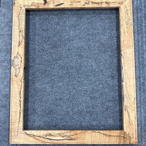 May include: A rectangular picture frame crafted from light-colored wood featuring prominent natural grain patterns and dark, irregular spalted markings throughout the surface. The frame has mitered corners and a smooth, flat profile design.
