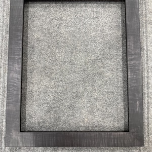 May include: A rectangular picture frame with a dark charcoal gray finish and a visible wood grain texture. The frame features a flat, wide border design with clean, sharp edges and a hollow center, providing a simple structure for displaying art.