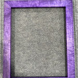 Pode incluir: Uma moldura quadrada de madeira com um vibrante acabamento roxo. A moldura apresenta uma textura de grão de madeira visível, adicionando um elemento natural ao seu design. Projetada para conter uma fotografia ou obra de arte.