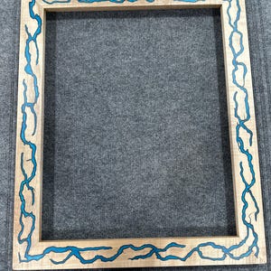 Pode incluir: Uma moldura retangular de madeira com acabamento castanho claro. A moldura é decorada com detalhes em resina azul turquesa que se assemelham a raios. A moldura está vazia, pronta para exibir uma fotografia ou obra de arte.