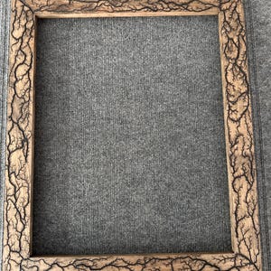 Pode incluir: Uma moldura quadrada de madeira com acabamento em madeira natural. As bordas da moldura apresentam um padrão único, escuro, semelhante a um raio. A moldura está vazia, pronta para exibir uma fotografia ou obra de arte.