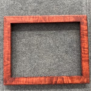 Könnte beinhalten: Ein rechteckiger Bilderrahmen aus Holz in einem satten, rotbraunen Farbton. Der Rahmen weist eine sichtbare Holzmaserung auf, die ihm Textur und visuelles Interesse verleiht. Der Rahmen ist leer und bereit für ein Foto oder Kunstwerk.