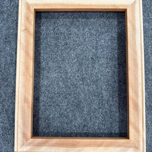 May include: A rectangular, unfinished wooden picture frame. The frame is made of light-colored wood with a natural grain. The frame has a simple, classic design, with a flat surface and a slightly raised edge. The frame is empty.