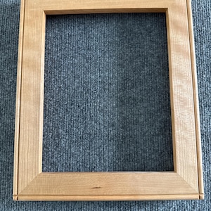 May include: A rectangular wooden picture frame with a light brown finish. The frame has a simple, classic design with a flat profile and a recessed inner edge. The wood grain is visible, adding texture to the frame.
