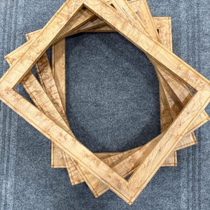 May include: A stack of three rectangular wooden picture frames featuring a light tan color with a distinct, swirling natural wood grain pattern. The frames are arranged in an overlapping, angled formation, creating a geometric layered effect.