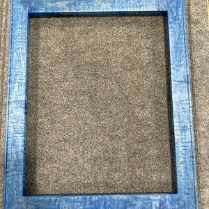 Puede incluir: Un marco de fotos de madera azul con una superficie lisa y ligeramente texturizada. El marco está vacío y listo para mostrar una foto o una obra de arte.