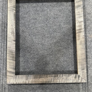 Pode incluir: Uma moldura de madeira cinza com uma superfície lisa e ligeiramente texturizada. A moldura está vazia e pronta para conter uma foto ou uma obra de arte.