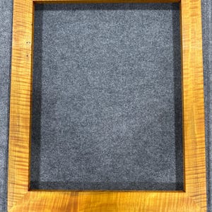 Pode incluir: Uma moldura de imagem de madeira natural com um acabamento quente de cor mel. A moldura tem um design simples e retangular e é feita de madeira maciça.