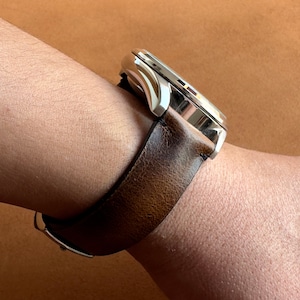 Op de afbeelding: Een zilveren polshorloge met een bruin leren bandje. Het horloge wordt gedragen aan de pols van een persoon.