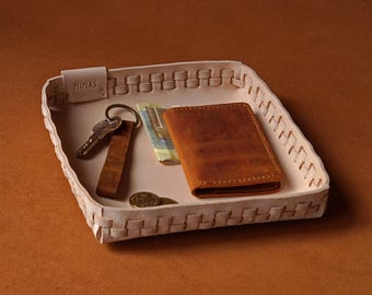 May include: A woven leather tray with a wallet, keys, and currency. The tray is off-white with a woven design around the edges. A brown leather wallet and key fob are inside the tray. The word "MINAS" is visible on the tray.