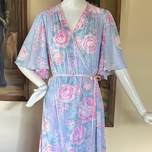 May include: A vintage, floor-length wrap dress in a light blue hue, adorned with a floral pattern of pink roses and green leaves. The dress features a V-neckline, flowing sleeves, and a matching pink tie belt. The dress is displayed on a mannequin.