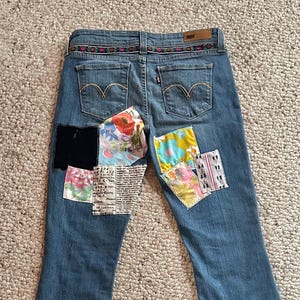 May include: Light blue denim jeans with patches of various colors and patterns sewn onto the back. The patches include floral prints, black fabric, and newspaper-style text. The jeans have a decorative waistband.