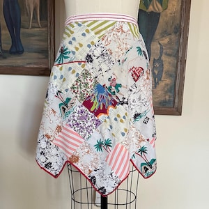 May include: A patchwork skirt with a variety of patterns, including floral, geometric, and tropical motifs. The skirt has a red trim and a striped waistband. The skirt is displayed on a mannequin.