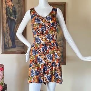 May include: A sleeveless, button-down romper with a V-neckline. The romper features a colorful abstract pattern in shades of orange, blue, yellow, and white. The garment is displayed on a white mannequin. The romper has a fitted bodice and a flared skirt.