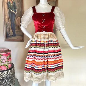 May include: A dress featuring a deep red velvet bodice with white lace-up detailing and sheer white puff sleeves. The skirt is cream-colored with horizontal bands of colorful woven patterns in orange, blue, green, and black, finished with a drawstring.