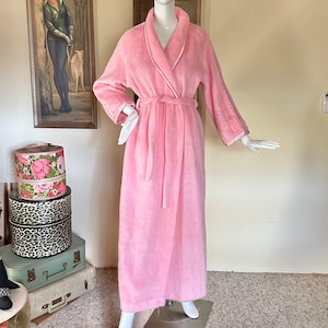 May include: A long, plush pink robe with a shawl collar and a matching belt. The robe has white trim along the collar, cuffs, and belt. The robe is displayed on a mannequin. The background includes vintage artwork and decorative hat boxes.