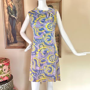 May include: A sleeveless knee-length dress featuring a vibrant, swirling abstract pattern in shades of yellow, lavender, and blue. The garment has a high neckline with a small decorative tie detail at the shoulder and a straight, fitted silhouette.