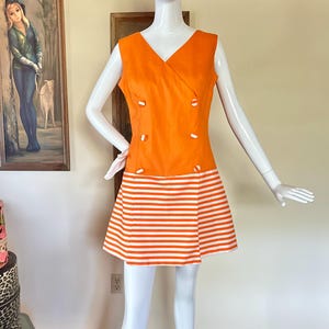 May include: A sleeveless orange dress featuring a V-neckline and a double-breasted front with six white buttons. The skirt portion of the dress displays a horizontal white and orange striped pattern, creating a distinct contrast with the solid orange bodice.