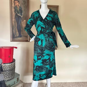 May include: A long-sleeved wrap dress in black and teal, featuring a floral pattern. The dress has a V-neck and a tie at the waist. The dress is displayed on a mannequin.