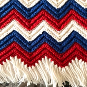 May include: A handmade crochet blanket with a chevron pattern in red, white, and blue. The blanket has a fringe of white yarn along the bottom edge. The blanket is a cozy home decor item.