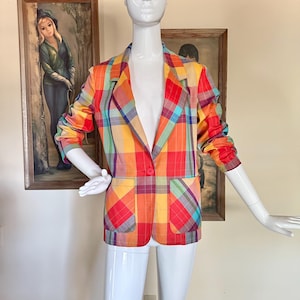 May include: A long-sleeve blazer featuring a vibrant plaid pattern in shades of red, orange, yellow, and teal. The garment includes a notched lapel, a single front button closure, and two large patch pockets on the lower front section of the jacket.